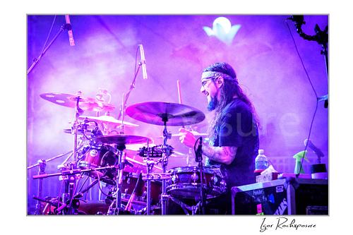 Horizontal color image of Mike Portnoy playing drums in side profile through purple stage smoke during a live performance