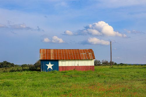A centered view of a rustic Texas Flag barn under a blue sky with soft white clouds in a green field.