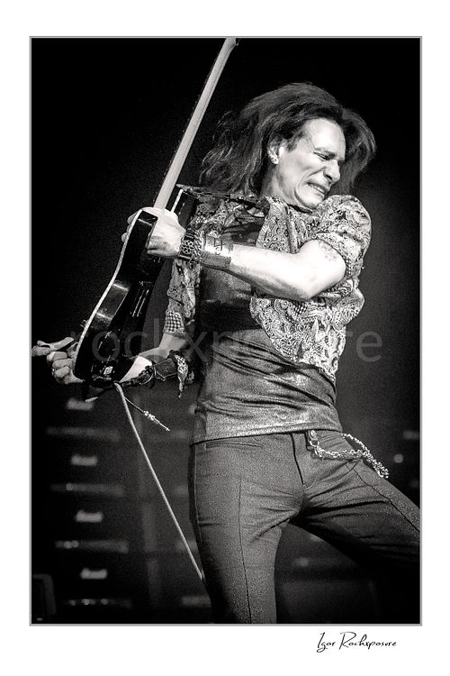 Vertical black and white image of Steve Vai swinging his guitar with flying hair and an intense expression in a dramatic stage pose