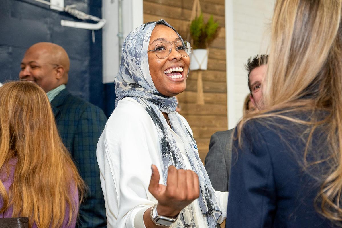 Corporate event photography capturing a professional woman in a patterned hijab smiling and engaging in conversation during a networking event, emphasizing authentic connection, representation, and inclusive professional community.