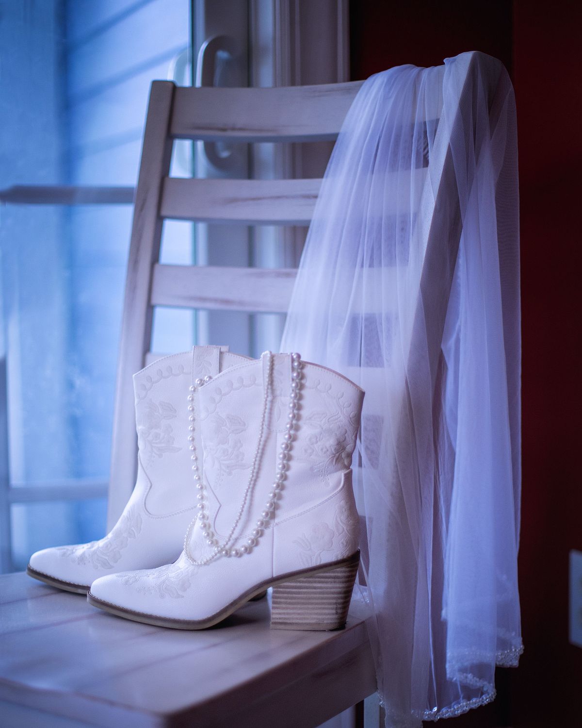 A cinematic detail shot of white bridal boots and a veil on a chair, illuminated by the cool, indigo tones of blue hour natural light.