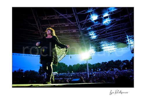 Horizontal color image of Ann Wilson walking on stage during a Heart concert, holding out her flowing stagewear before a large outdoor crowd at dusk under bright backlight