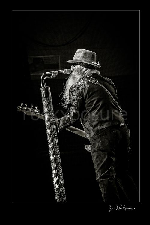 Vertical black and white image of Dusty Hill of ZZ Top performing live on stage with a bass guitar, singing beside a tall textured microphone stand in his signature hat, dark glasses, and long beard
