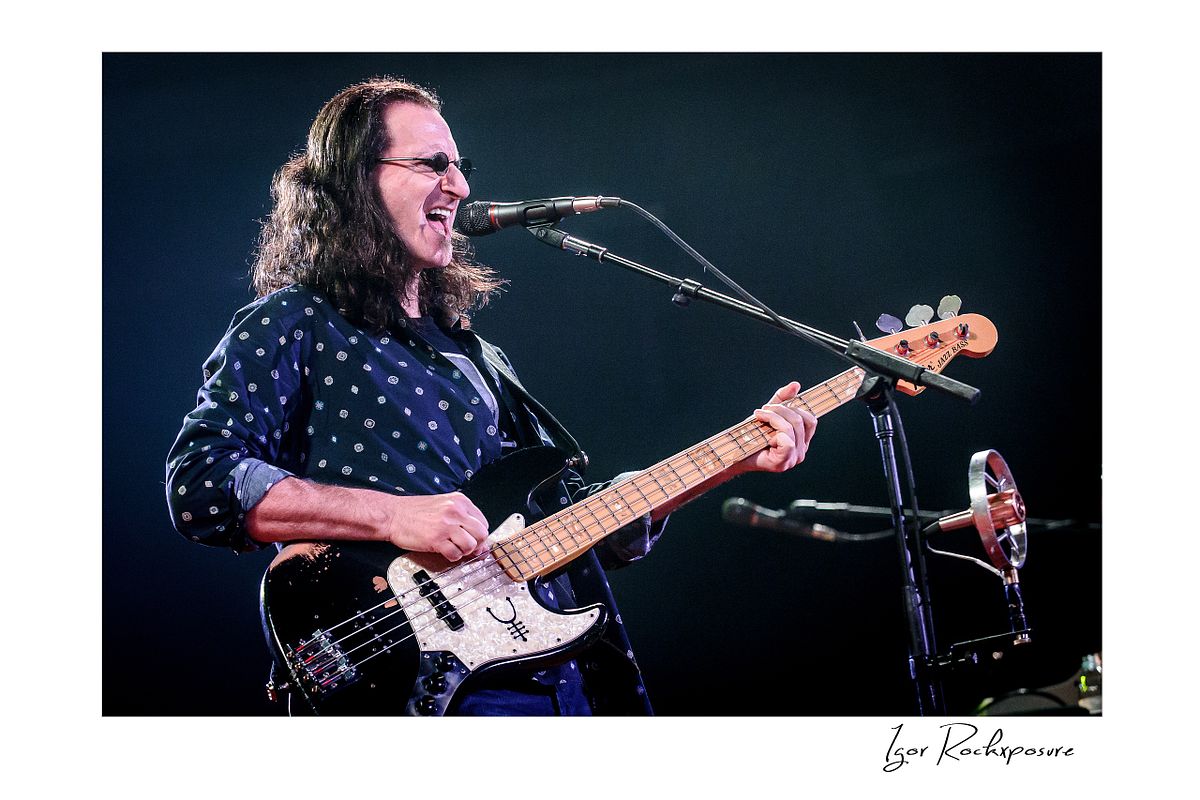 Horizontal concert photography of Geddy Lee playing bass guitar and singing into a microphone under stage lighting