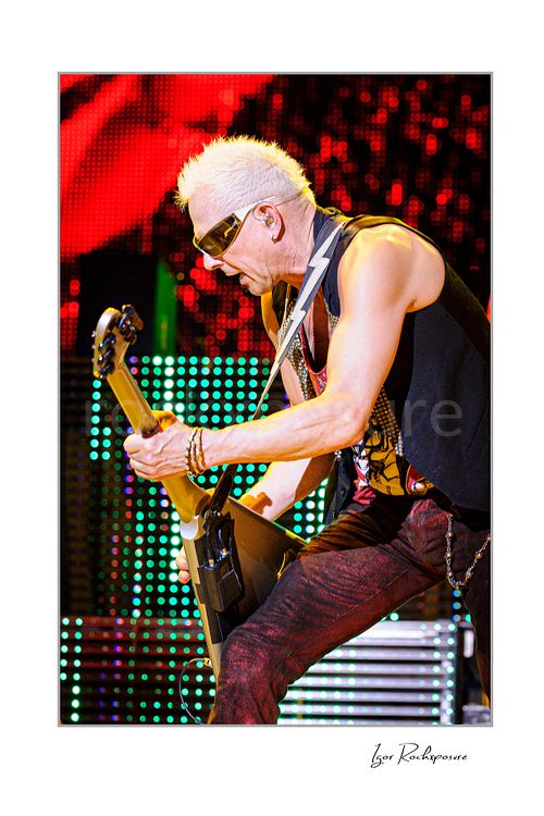 Vertical color image of Rudolf Schenker of Scorpions playing guitar live in sunglasses with red pants and glowing LED stage lights