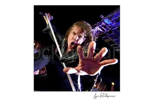 Square color image of Joey Tempest of Europe performing live on stage with a white microphone stand, black leather jacket, dramatic low-angle perspective, and outstretched hand reaching toward the camera