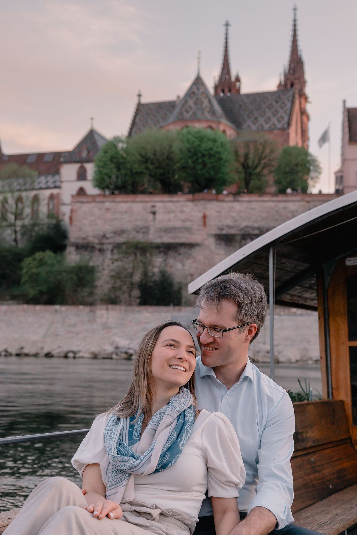 A smiling Basel couple sits in a boat, with the man embracing the woman. A historic cathedral with tall spires is visible in the background, creating a romantic atmosphere.