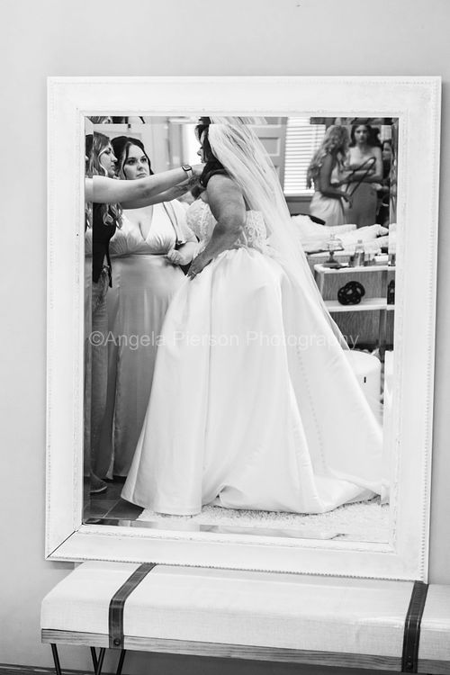 A bride's reflection is photographed through a mirror as bridesmaids assist her in getting ready