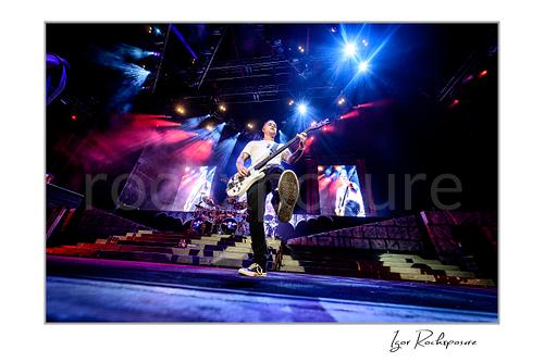 Horizontal color image of Johnny Christ of Avenged Sevenfold playing bass on stage in a dramatic low-angle wide shot with one foot raised and vivid blue and magenta concert lights