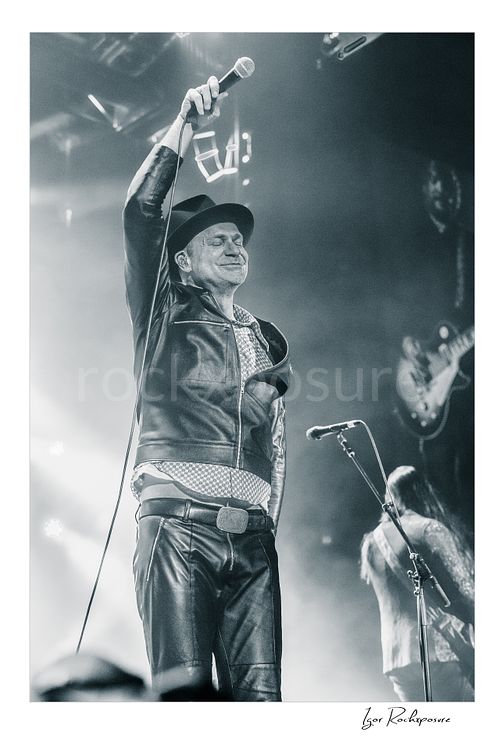 Vertical black and white image of Gord Downie of The Tragically Hip raising a microphone high above his head, wearing a hat and leather jacket in bright concert haze with a blurred band member behind him