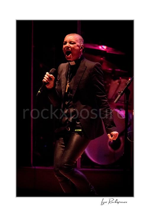 Vertical color image of Sinéad O’Connor singing into a microphone on stage with a drum kit behind her