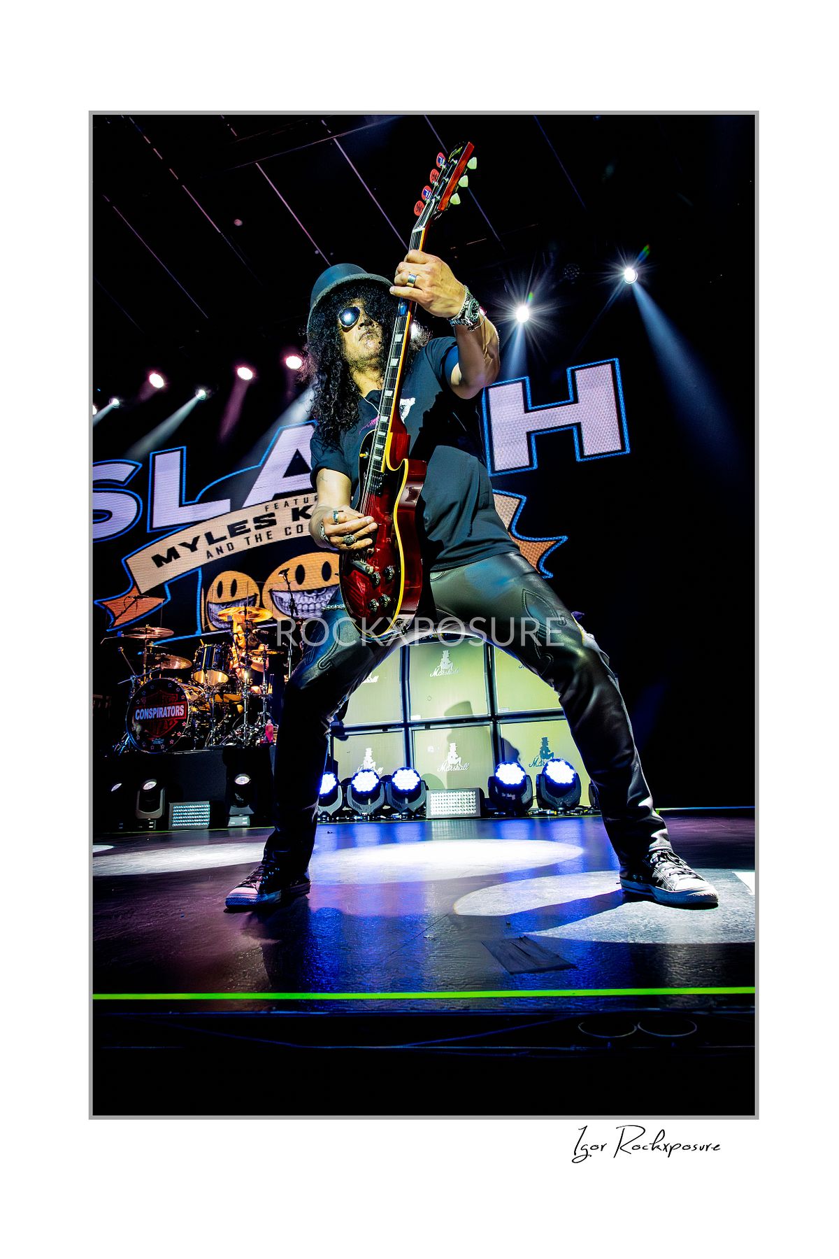 Vertical concert photography of Slash raising his electric guitar overhead in a wide power stance under colorful stage lighting