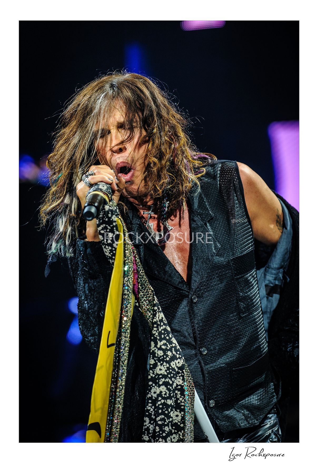 Vertical concert photography of Steven Tyler singing into a microphone with scarf-wrapped stand under dramatic blue and purple stage lighting