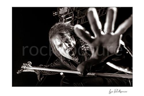 Horizontal black and white image of Joey Tempest of Europe performing live on stage with a white microphone stand, dramatic low-angle perspective, open mouth scream, and outstretched hand reaching toward the camera