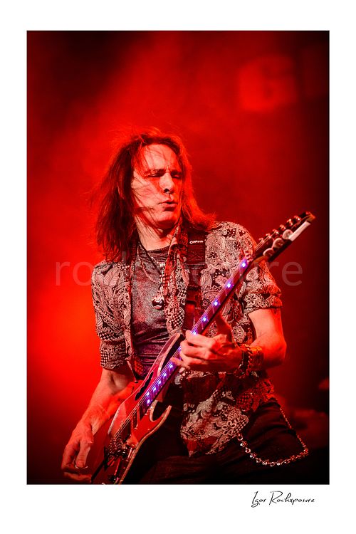 Vertical color image of Steve Vai playing guitar under vivid red stage lighting with glowing fret markers and windswept hair