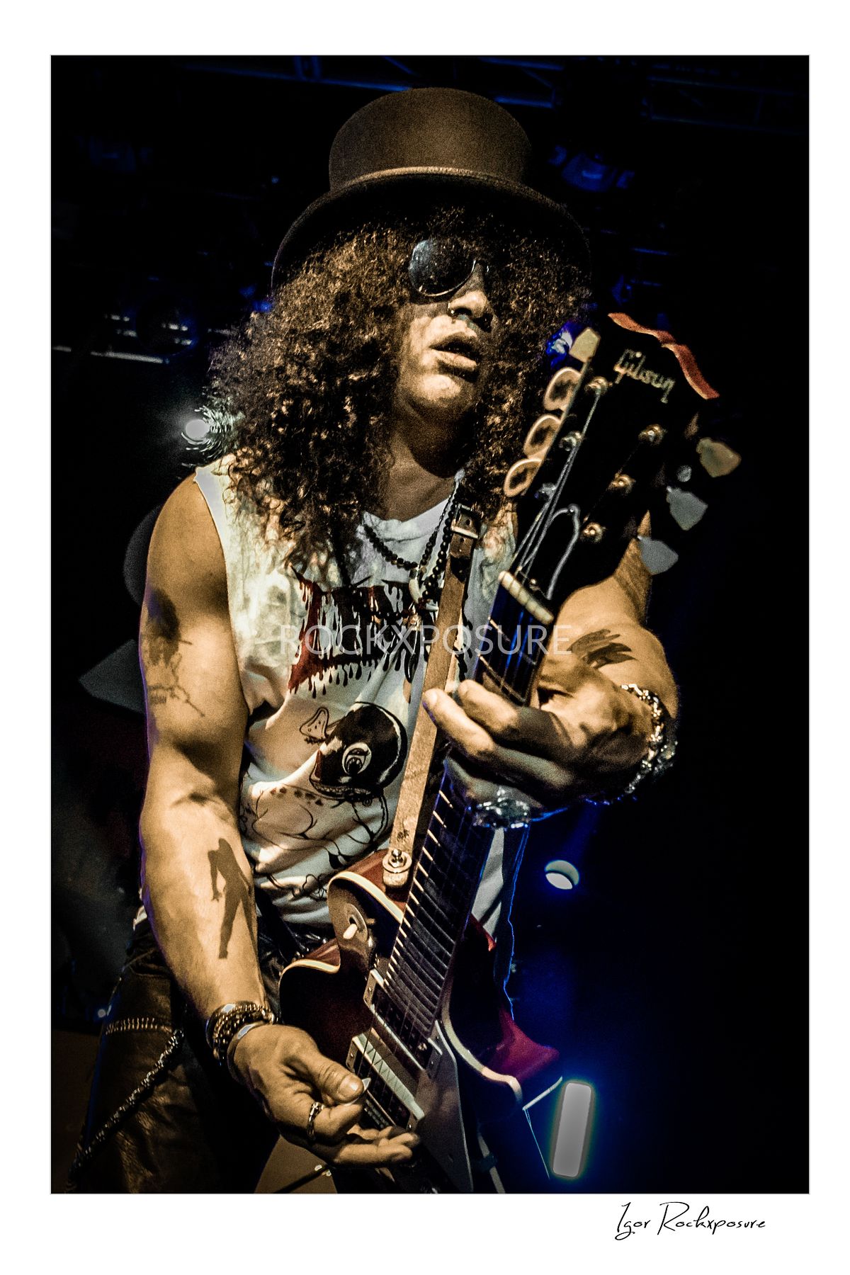 Vertical concert photography of Slash playing electric guitar wearing his signature top hat and sunglasses under dramatic stage lighting