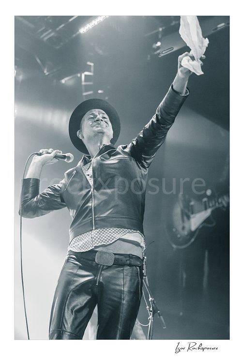 Vertical black and white image of Gord Downie of The Tragically Hip holding a microphone and lifting a white cloth high on stage, wearing a hat and leather jacket in bright concert haze