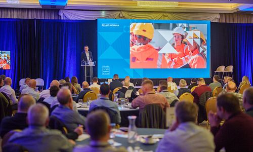 Wide shot of a corporate education event with a speaker on stage and a large audience.