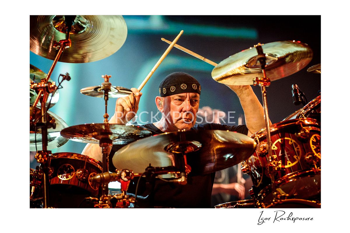 Horizontal concert photography of Neil Peart playing drums surrounded by a large drum kit under warm stage lighting
