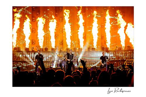 Horizontal color image of Nickelback performing onstage before a packed crowd with towering fire columns and bright concert beams