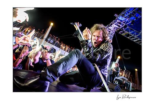 Horizontal color image of Joey Tempest of Europe performing live on stage in a black leather jacket with a white microphone stand, crouched pose, and fans photographing him at the stage edge
