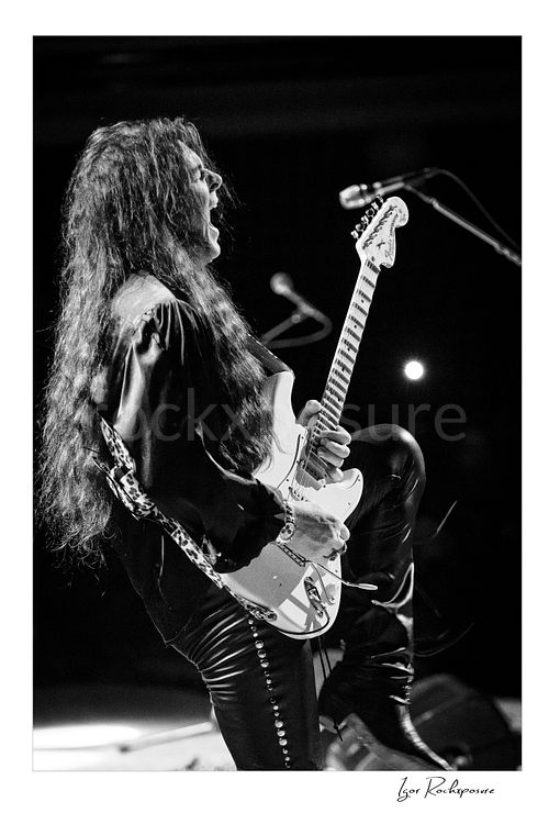 Vertical black and white image of Yngwie Malmsteen performing live with a white guitar, long hair flowing and one knee raised in a dramatic stage pose beside the microphone