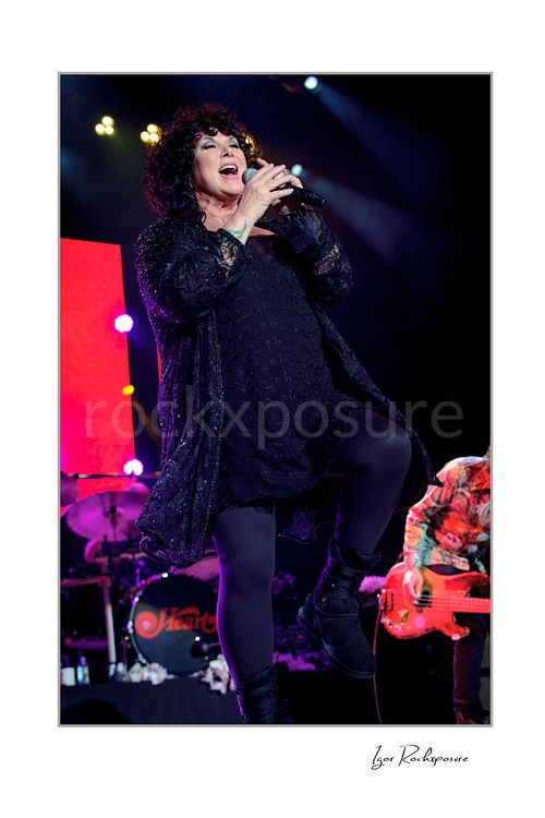 Vertical color image of Ann Wilson singing into a microphone during a Heart concert, lifting one leg in a dynamic stage pose under vivid purple and blue light