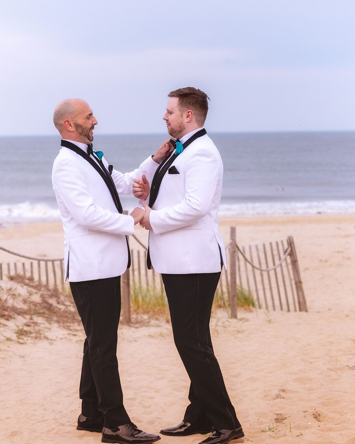 Two grooms in white tuxedos sharing an intimate moment on a beach, an example of fine art wedding photography with natural ocean light.