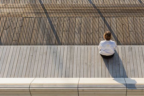 Minimalist portrait of a woman sitting alone on a wooden deck in sunlight, photographed from above in Stockholm by Mats Karlsson.