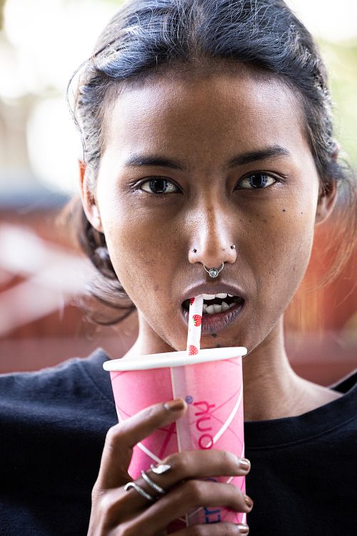 A headshot of an Indian woman, holding a disposable coffee cup with a straw, dressed in a black t-shirt, and adorned with earrings and a nose ring.