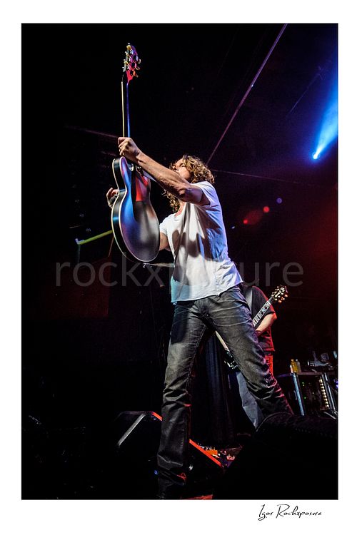 Vertical color image of Chris Cornell performing live on stage, holding his guitar upright under dramatic blue and red concert lighting