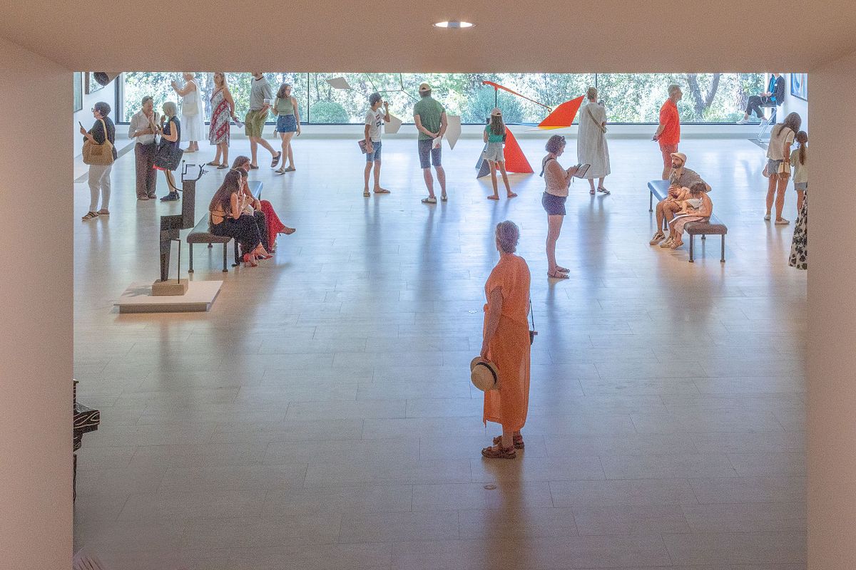 Bes&ouml;kare i en ljus konsthall med skulpturer och installationer; en kvinna i orange st&aring;r mitt i rummet.