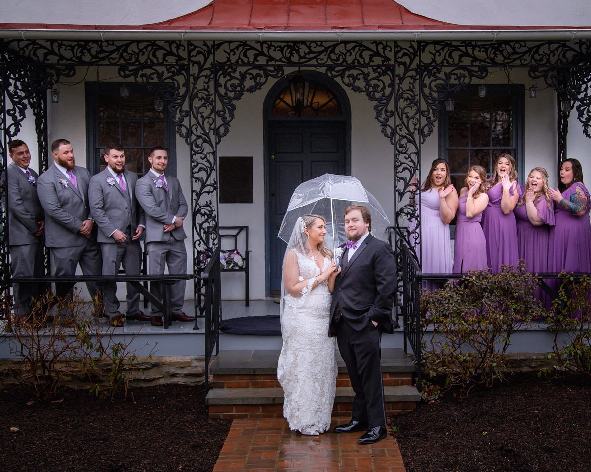 bride and groom posing in front of a stone house under an umbrella. The wedding party is under the covered porch cheering them on