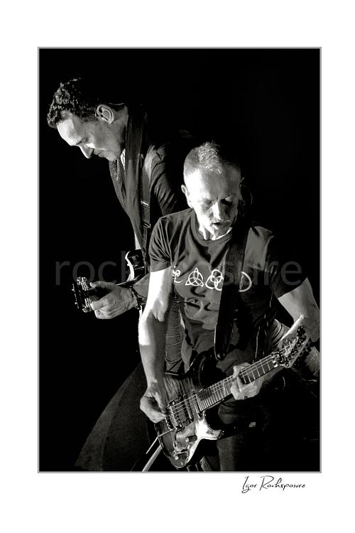Vertical black and white image of Vivian Campbell and Phil Collen of Def Leppard performing live on stage, with Campbell at left and Collen at right playing guitar against a dark background