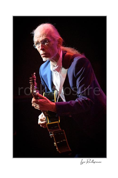 Vertical color image of Steve Howe of Yes playing guitar onstage under warm red concert light