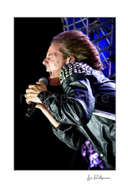 Vertical color image of Joey Tempest of Europe performing live on stage with a handheld microphone, black leather jacket, windblown hair, and blue stage truss lighting