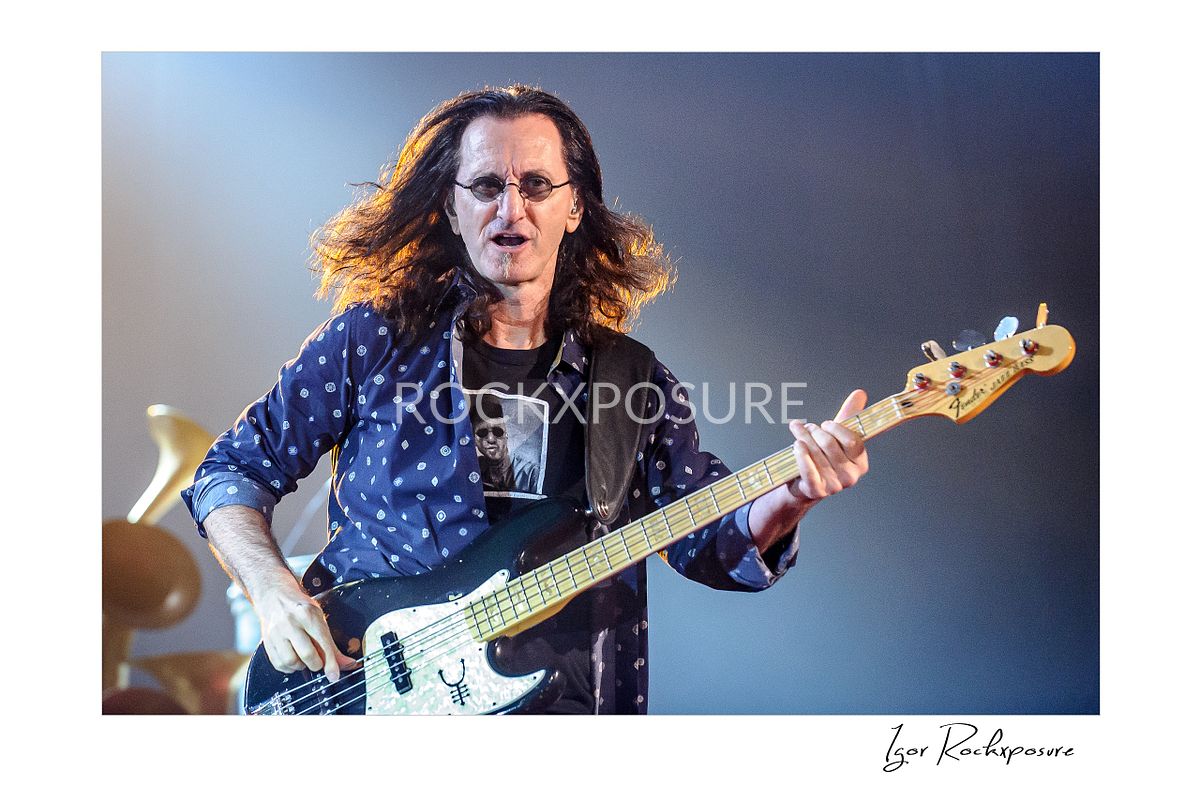 Horizontal concert photography of Geddy Lee playing bass guitar under soft stage lighting