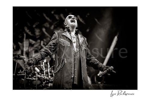 Horizontal black and white image of Rob Halford of Judas Priest performing live on stage, holding a microphone with his arms extended and the drum kit visible in the background