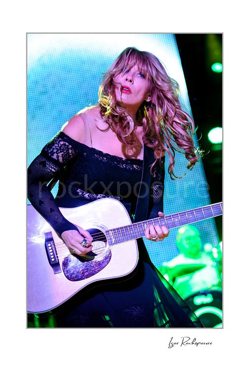 Vertical color image of Nancy Wilson performing with acoustic guitar during a Heart concert, with flowing hair and vivid green stage light