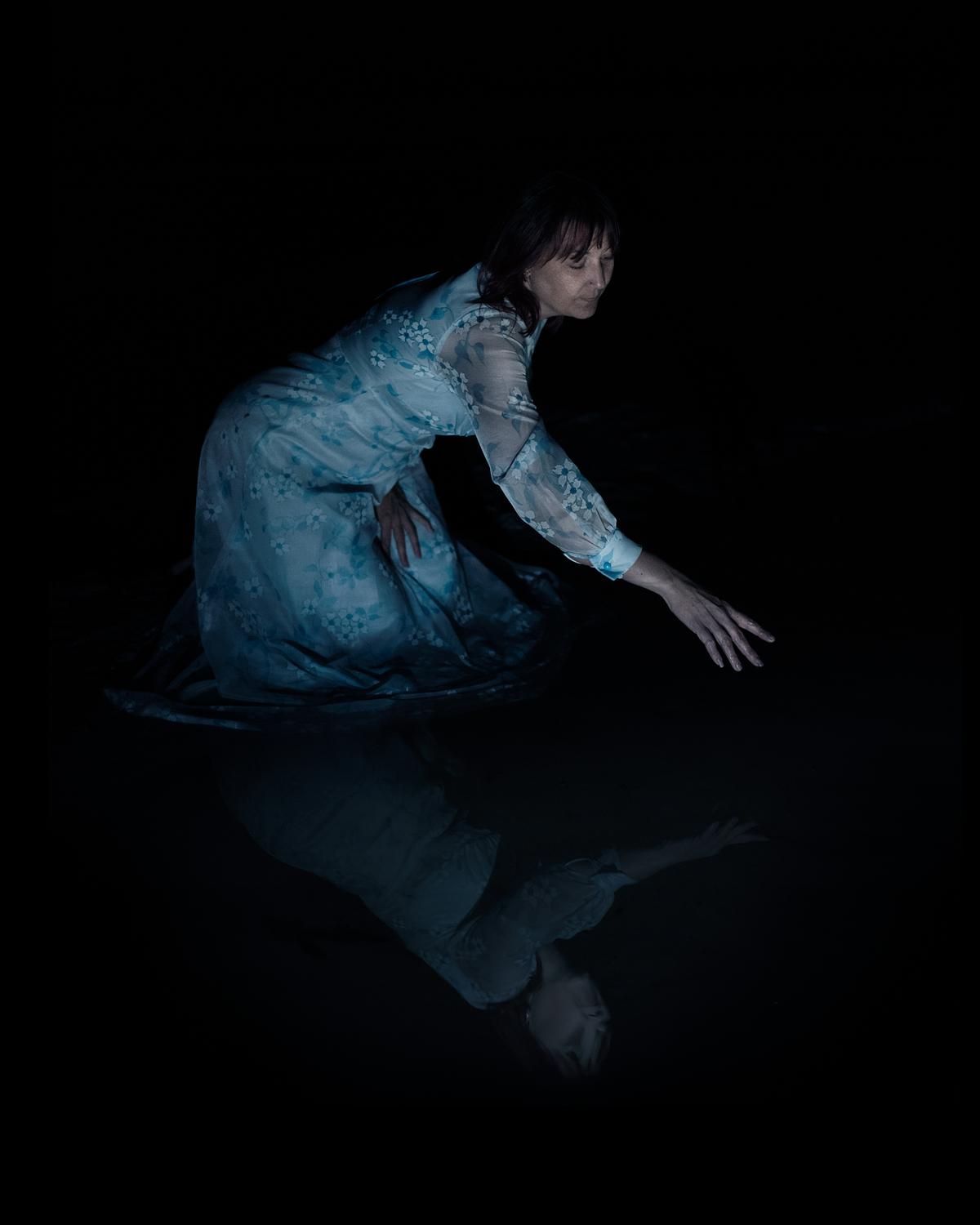 A woman reaching for her distorted reflection in the water.