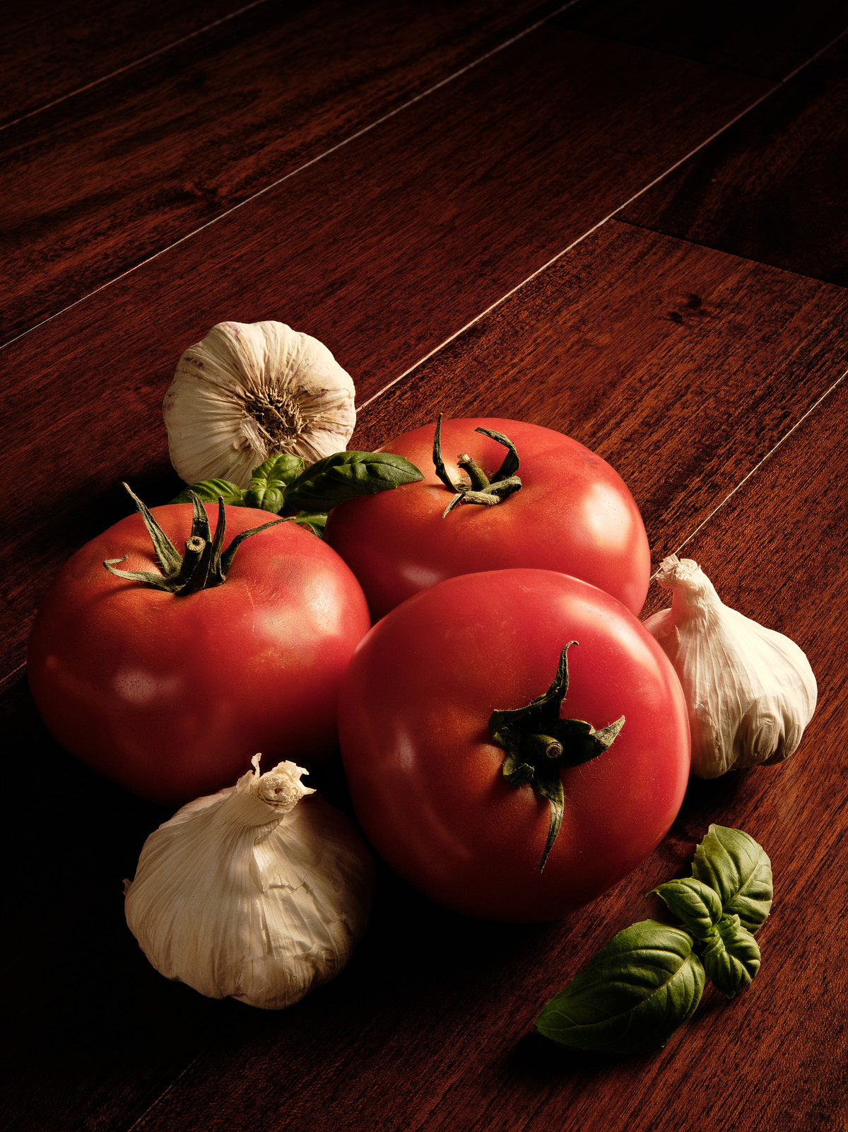 Three red tomatoes with garlic and basil on mahogany wood.
