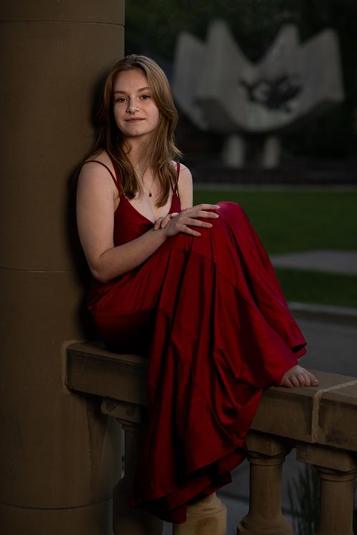 Grad photo of a young woman sitting on concrete railing with a unique sculpture in the background.