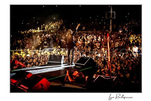 Horizontal color image of C.C. DeVille walking out on stage during a Poison concert toward a packed crowd with raised arm under warm concert lighting
