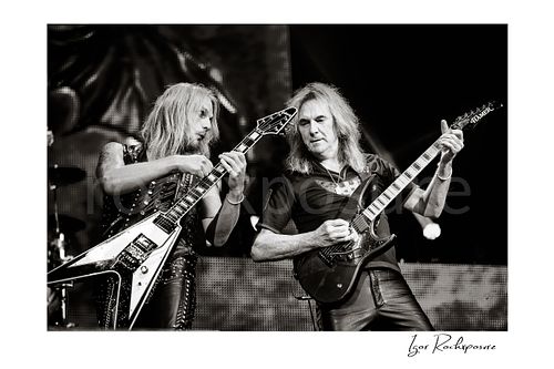 Horizontal black and white image of Richie Faulkner and Glenn Tipton of Judas Priest performing live on stage, with Faulkner at left playing flying V guitar and Tipton at right playing electric guitar