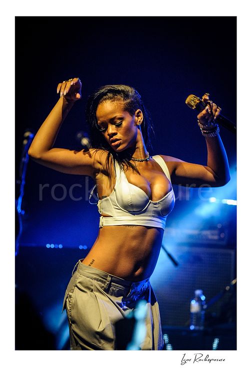 Vertical color image of Rihanna dancing on stage with a gold microphone, white top, and deep blue concert lighting