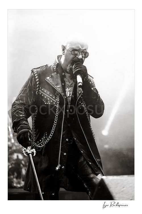 Vertical black and white image of Rob Halford of Judas Priest performing live on stage, singing into a microphone while wearing a studded leather coat and sunglasses