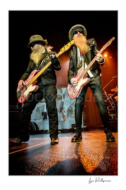 Vertical color image of Dusty Hill and Billy Gibbons of ZZ Top performing live on stage with matching red guitars, wearing signature hats, dark glasses, and long beards under warm concert light