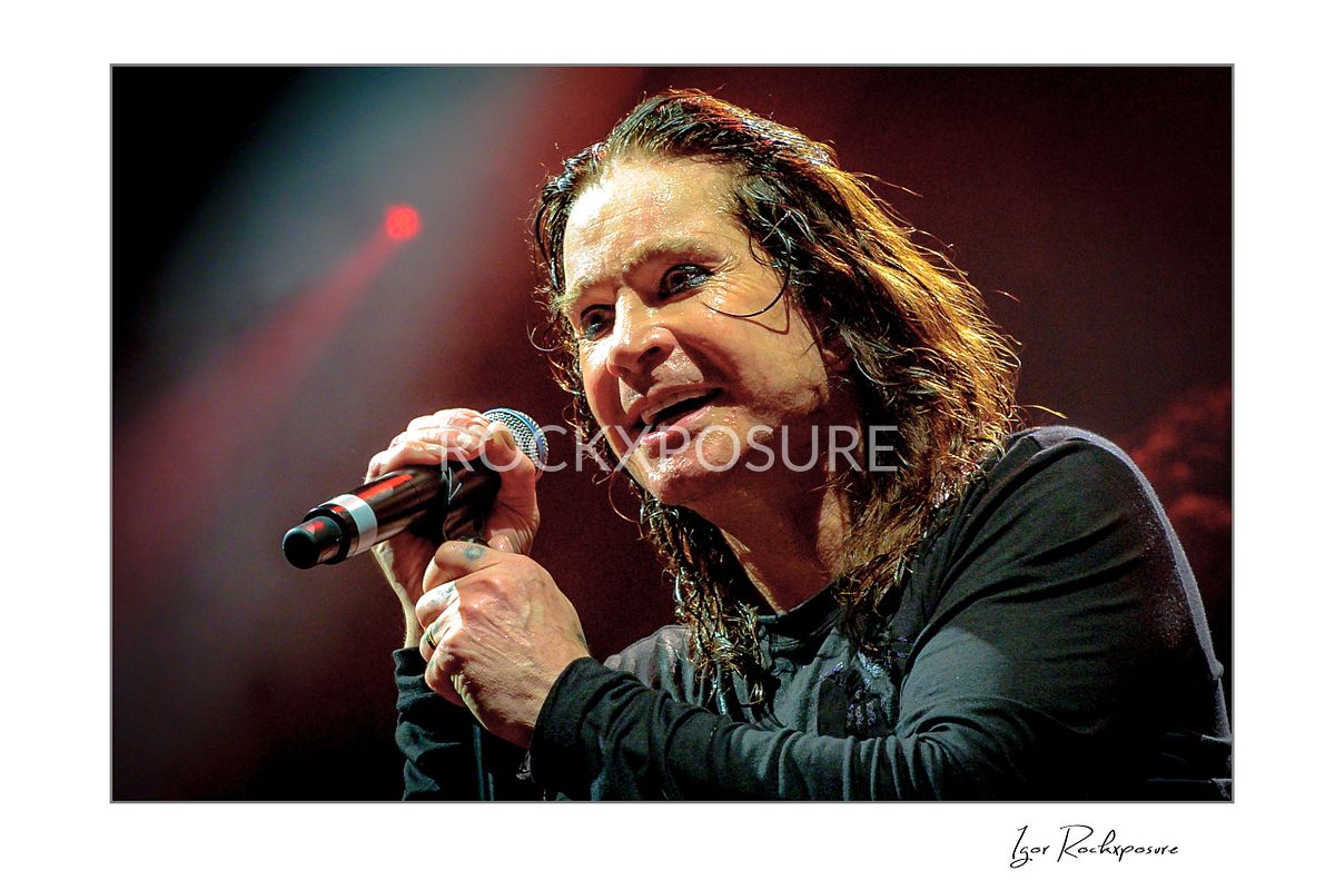 Ozzy Osbourne of Black Sabbath singing into a handheld microphone during a live performance under warm red concert lighting