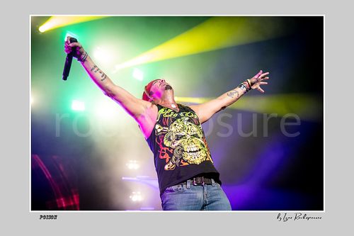 Horizontal color image of Bret Michaels performing live during a Poison concert, raising both arms with microphone in hand under vivid yellow and purple stage lights