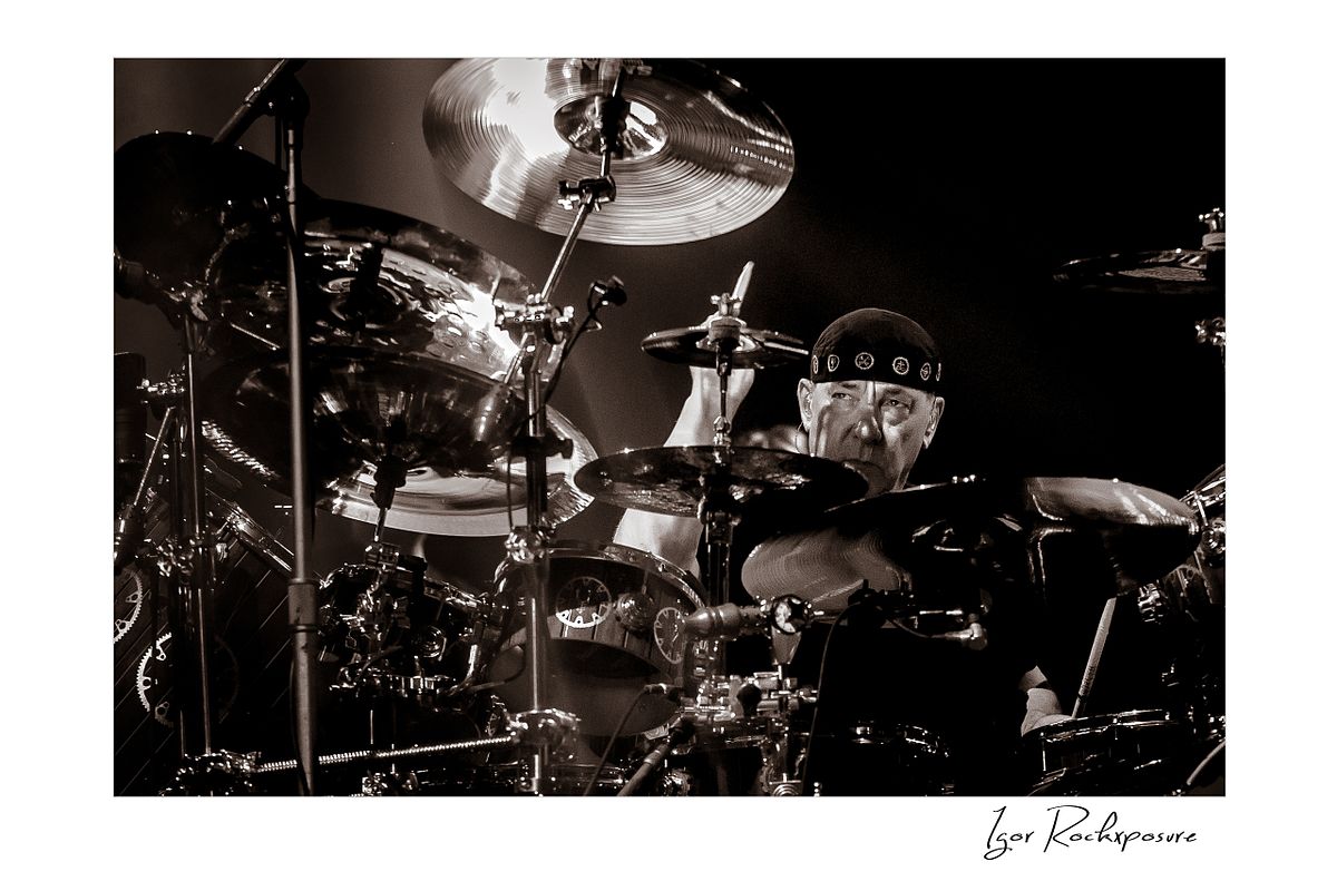 Horizontal concert photography of Neil Peart playing a large drum kit in black and white photography under stage lighting